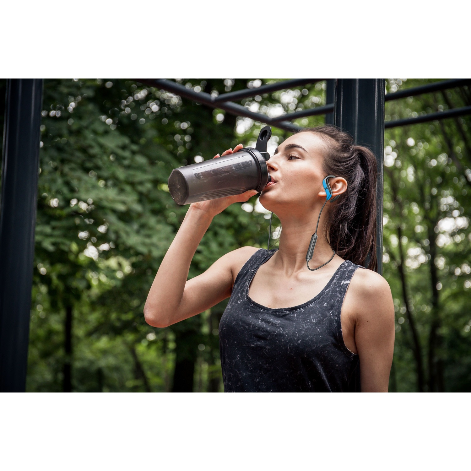 Una donna beve da uno shaker grigio, indossa cuffie blu, si allena all'aperto.
