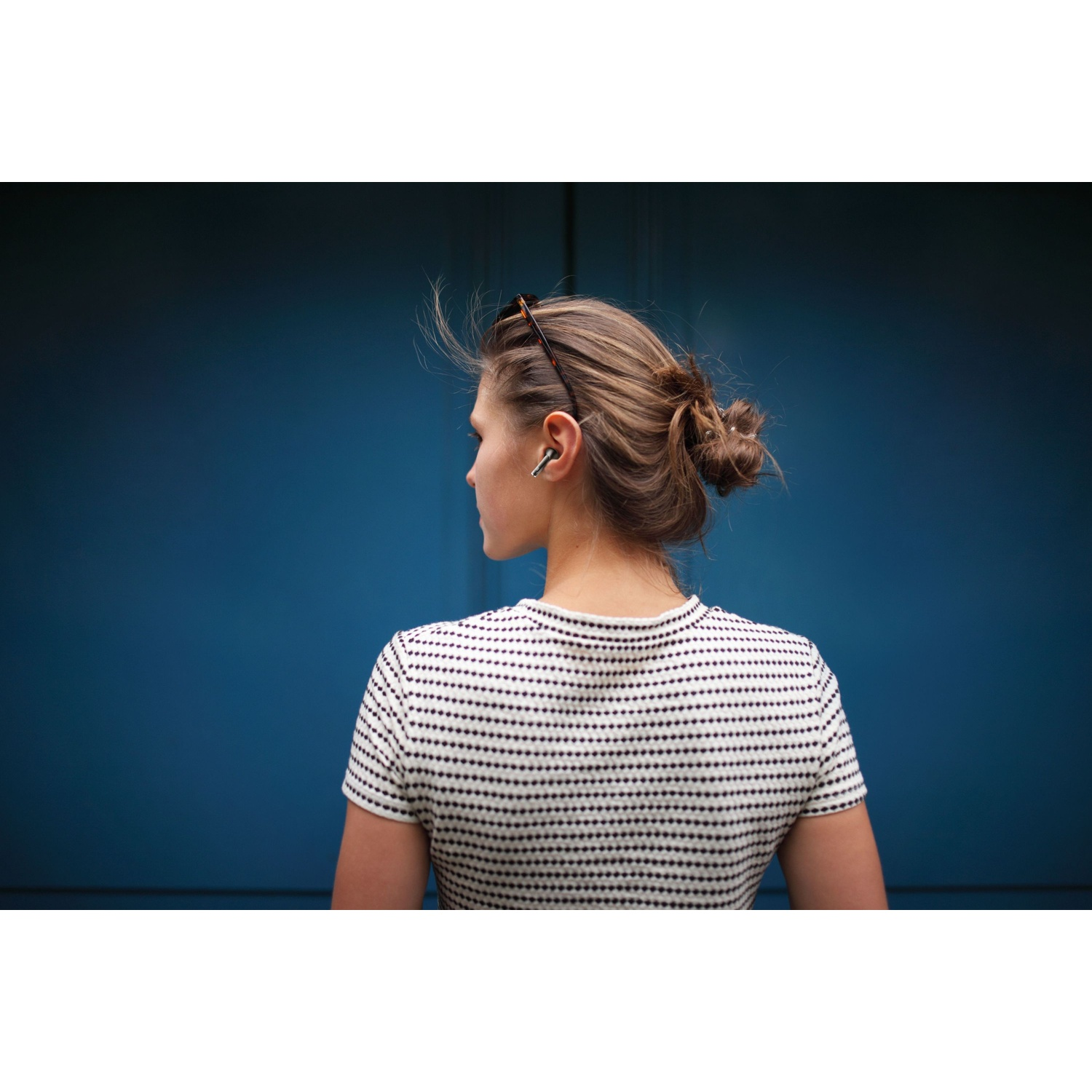 Donna con auricolari rivolta a sinistra su sfondo blu, vista da dietro.