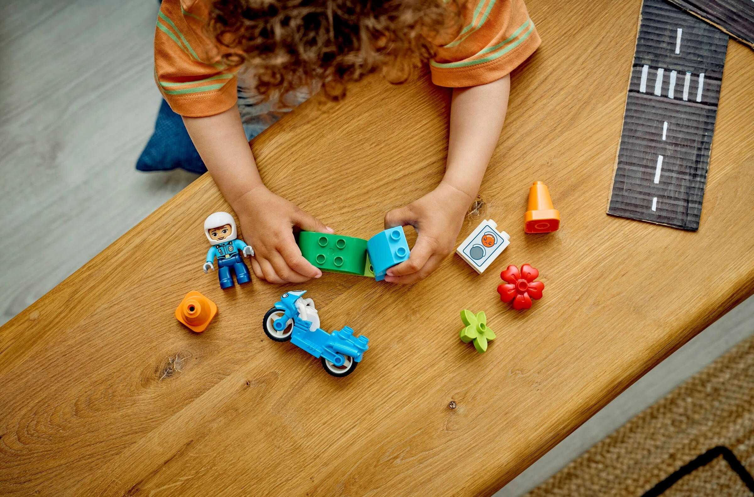 Dziecko buduje z kolorowych klocków na drewnianym stole. Zawiera figurki, motocykl, pachołki.