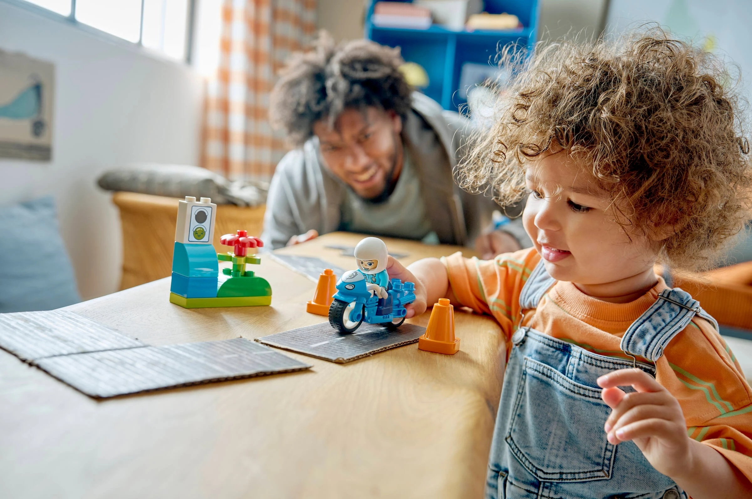 Dziecko bawi się niebieskim motocyklem zabawką na drewnianym stole, mężczyzna śmieje się w tle.