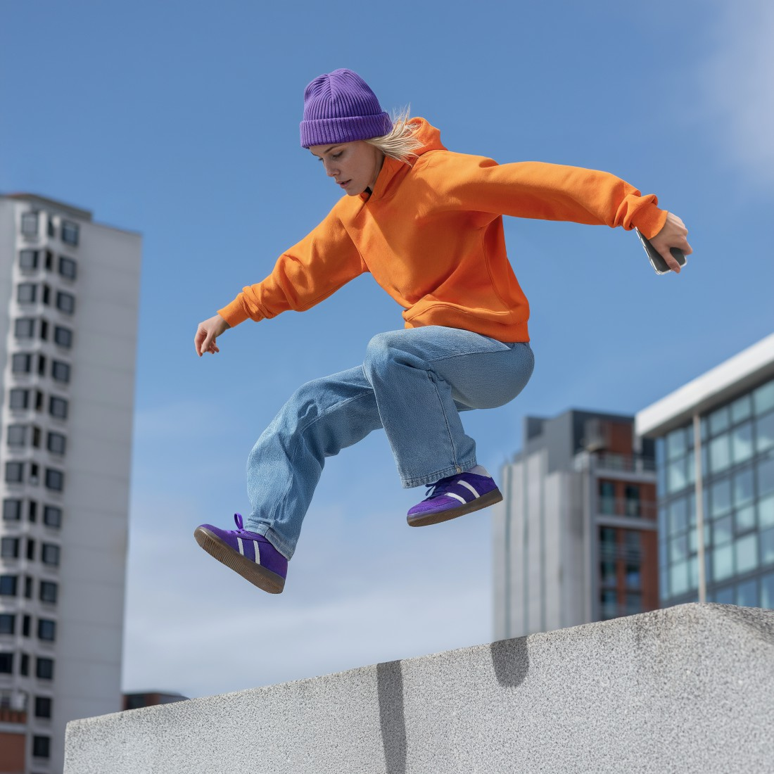 Junge Person in Orange Hoodie springt über Mauer, lila Mütze und Schuhe.