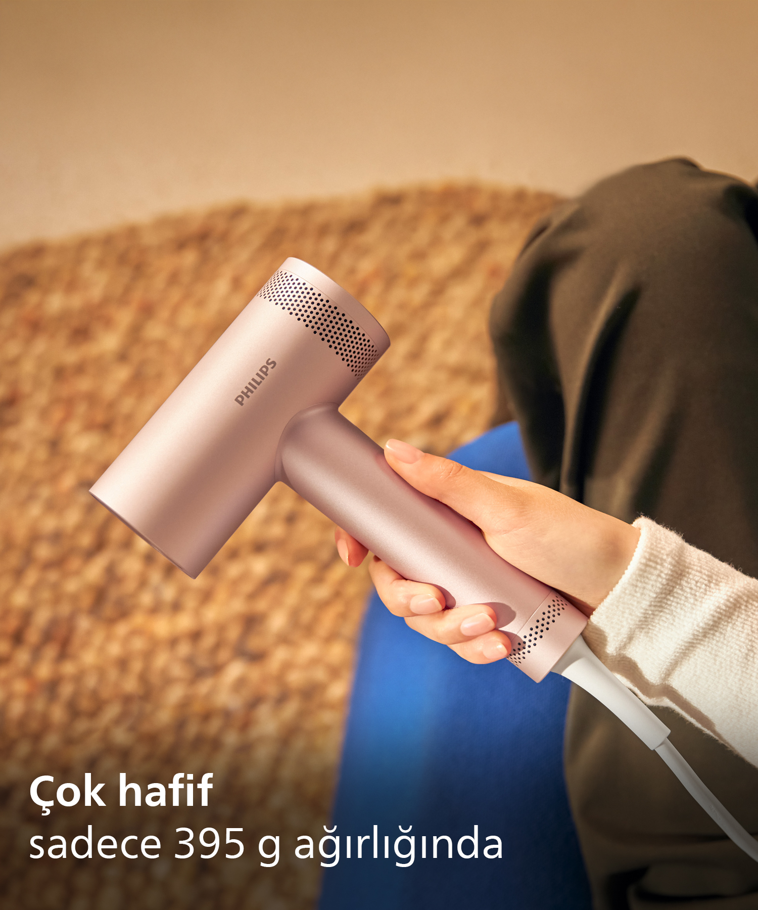 Bir kişi hafif, roz altın rengi Philips saç kurutma makinesi tutuyor. Arkada halı var.
