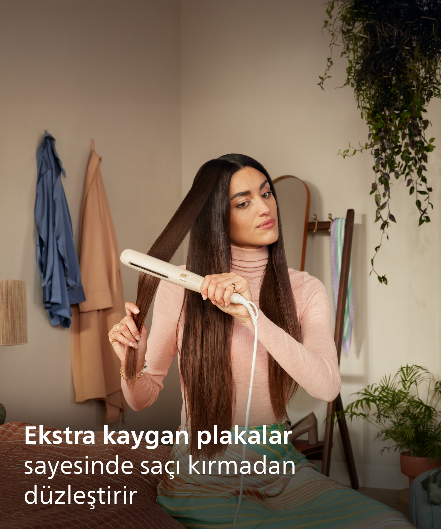 Kadın, uzun kahverengi saçlarını beyaz düzleştiriciyle düzleştiriyor. Pembe bluz giyiyor.