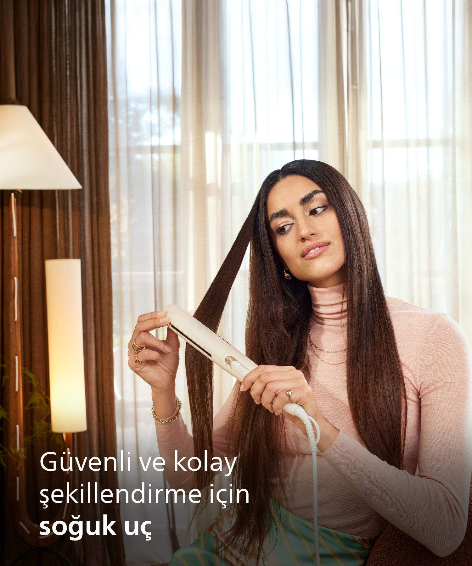 Kadın, rahat bir odada beyaz düzleştirici ile uzun kahverengi saçlarını şekillendiriyor.