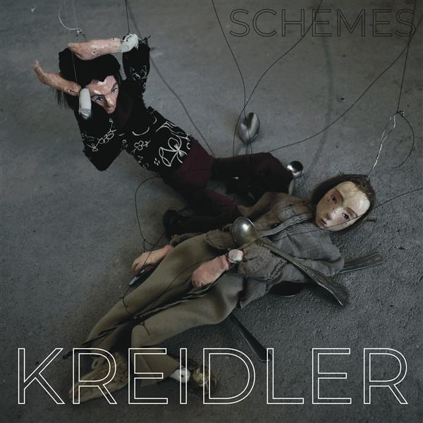 Zwei Puppen liegen nebeneinander auf Beton, verbunden mit Fäden. "SCHEMES" oben, "KREIDLER" unten.
