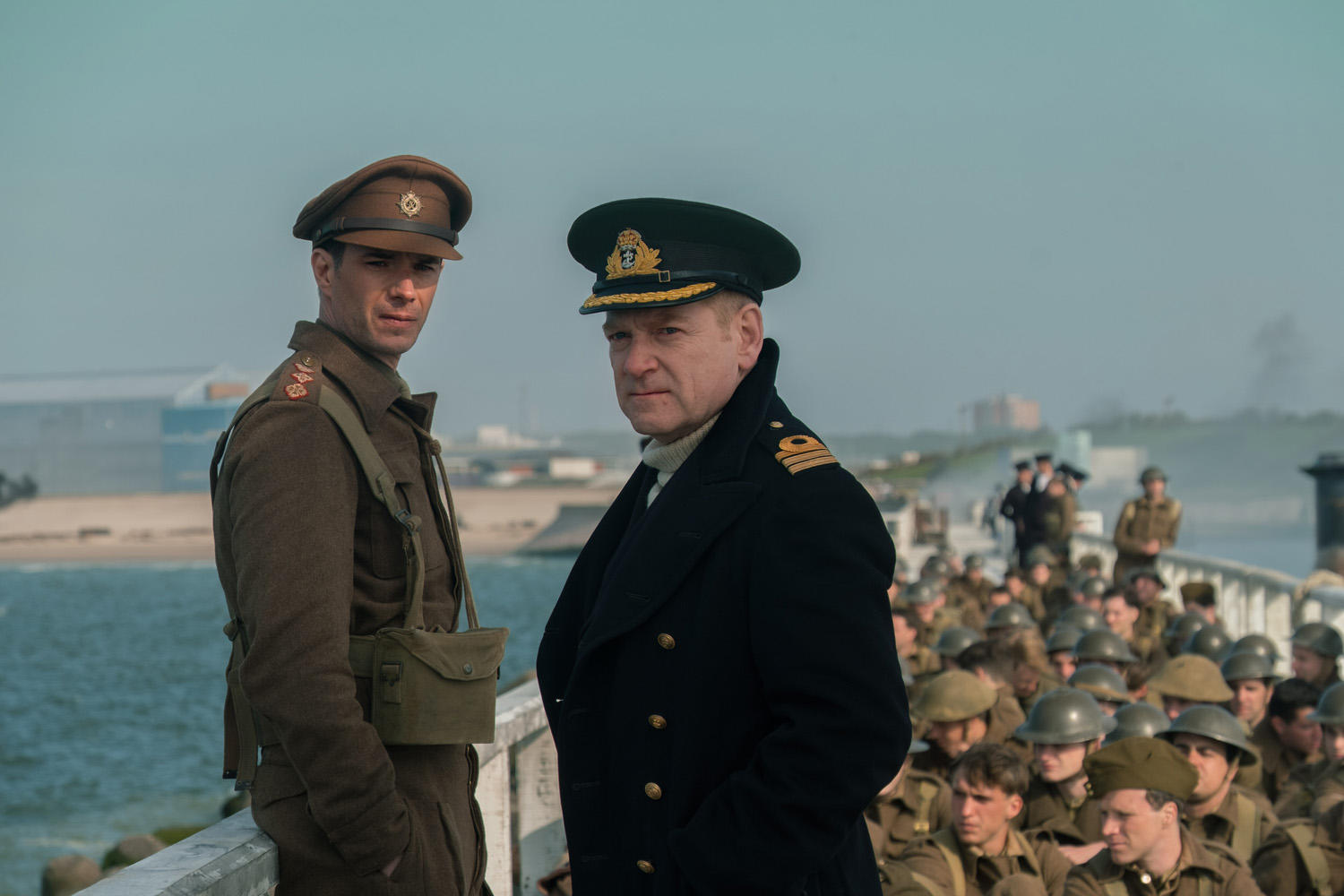 Zwei Männer in Uniform, einer in brauner Armeeuniform, der andere in blauer Marineuniform, stehen vor Soldaten.