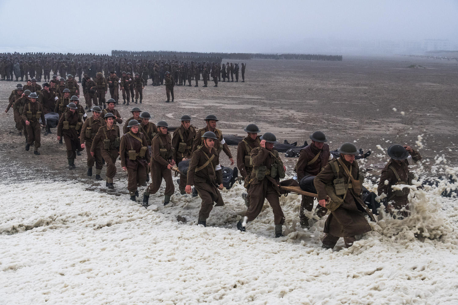 Soldaten in Uniform tragen Verwundete auf Tragbahren am Strand, während andere aufgereiht sind.