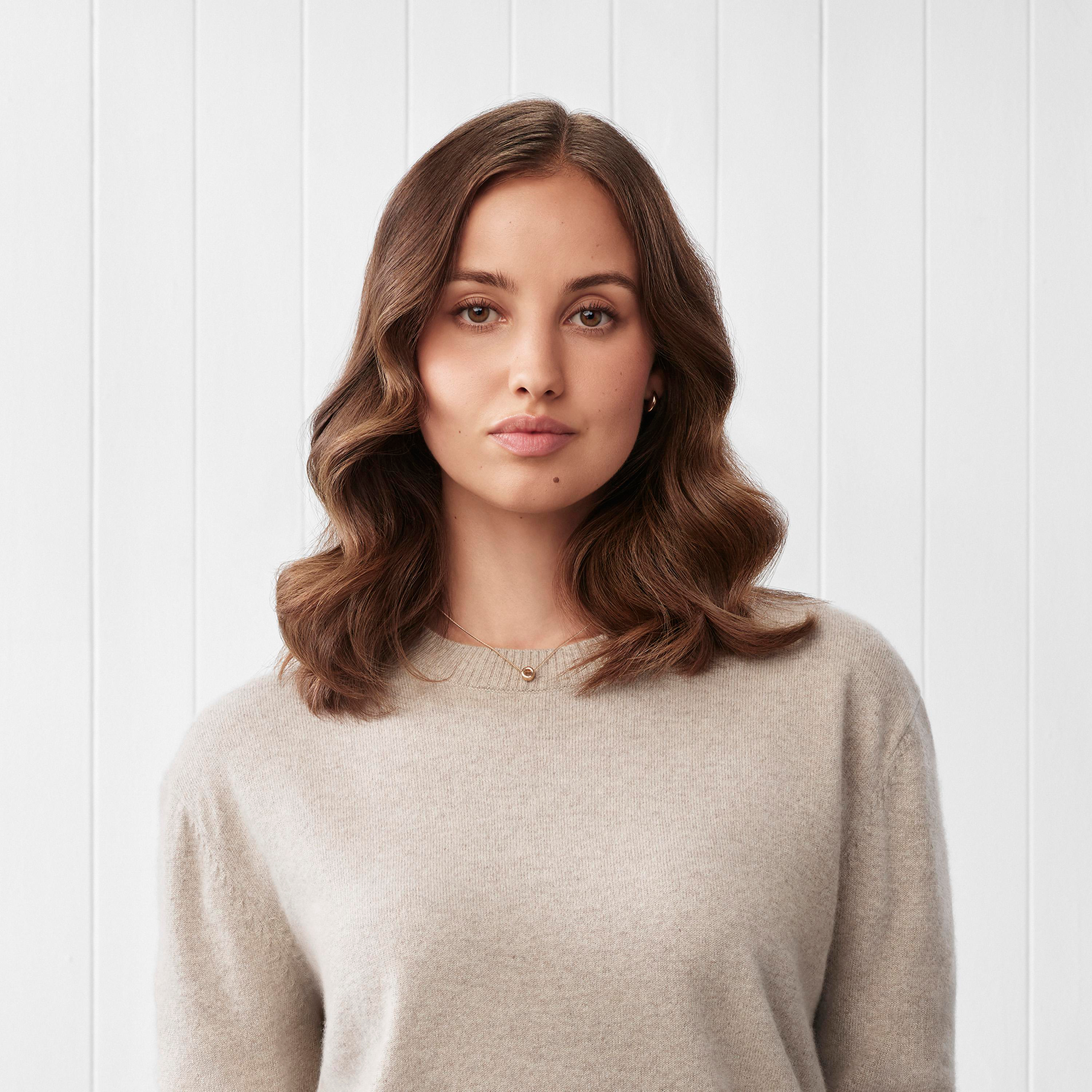 Eine Frau mit braunen Haaren trägt einen beigen Pullover vor weißem Hintergrund.