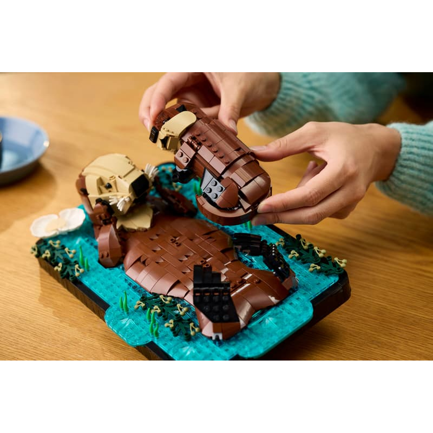 Hands building a brown Lego otter on a blue base with green plants.