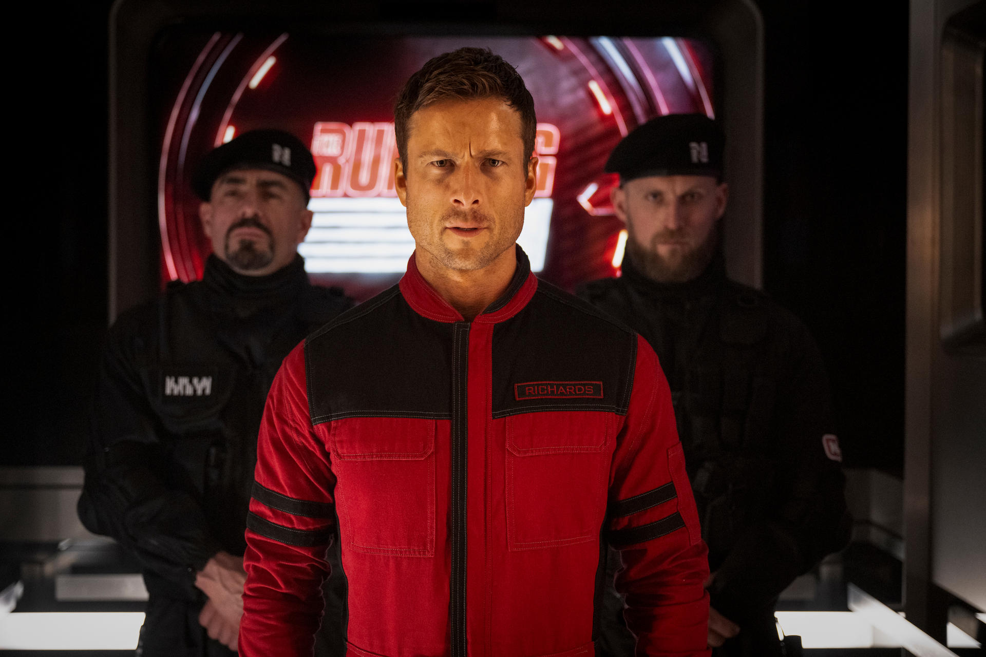 Ein Mann in Rot und Schwarz mit zwei Männern in Schwarz dahinter.