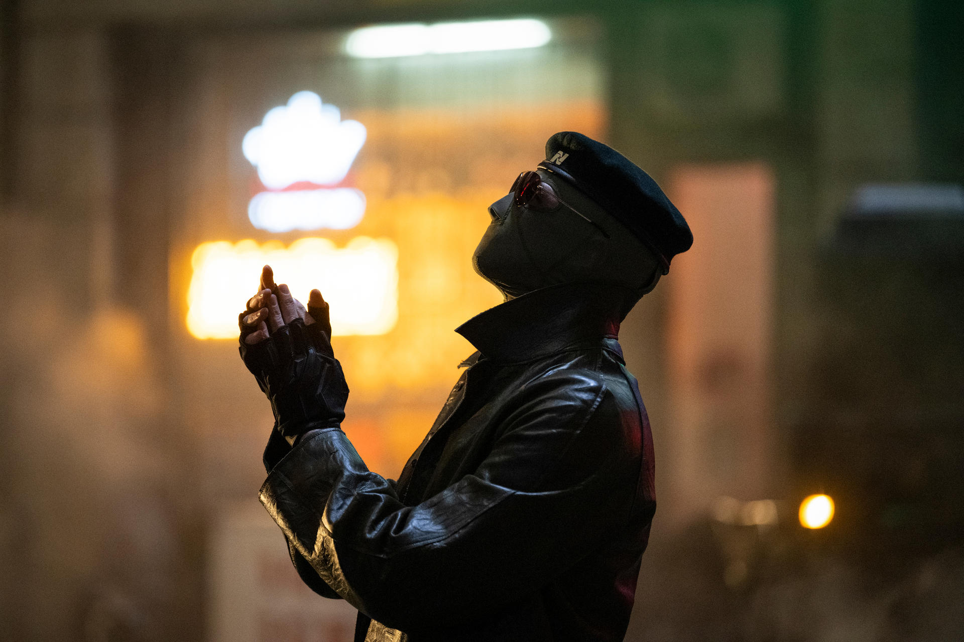Mann in Lederjacke und Maske mit Händen vor dem Gesicht, leuchtender Hintergrund.