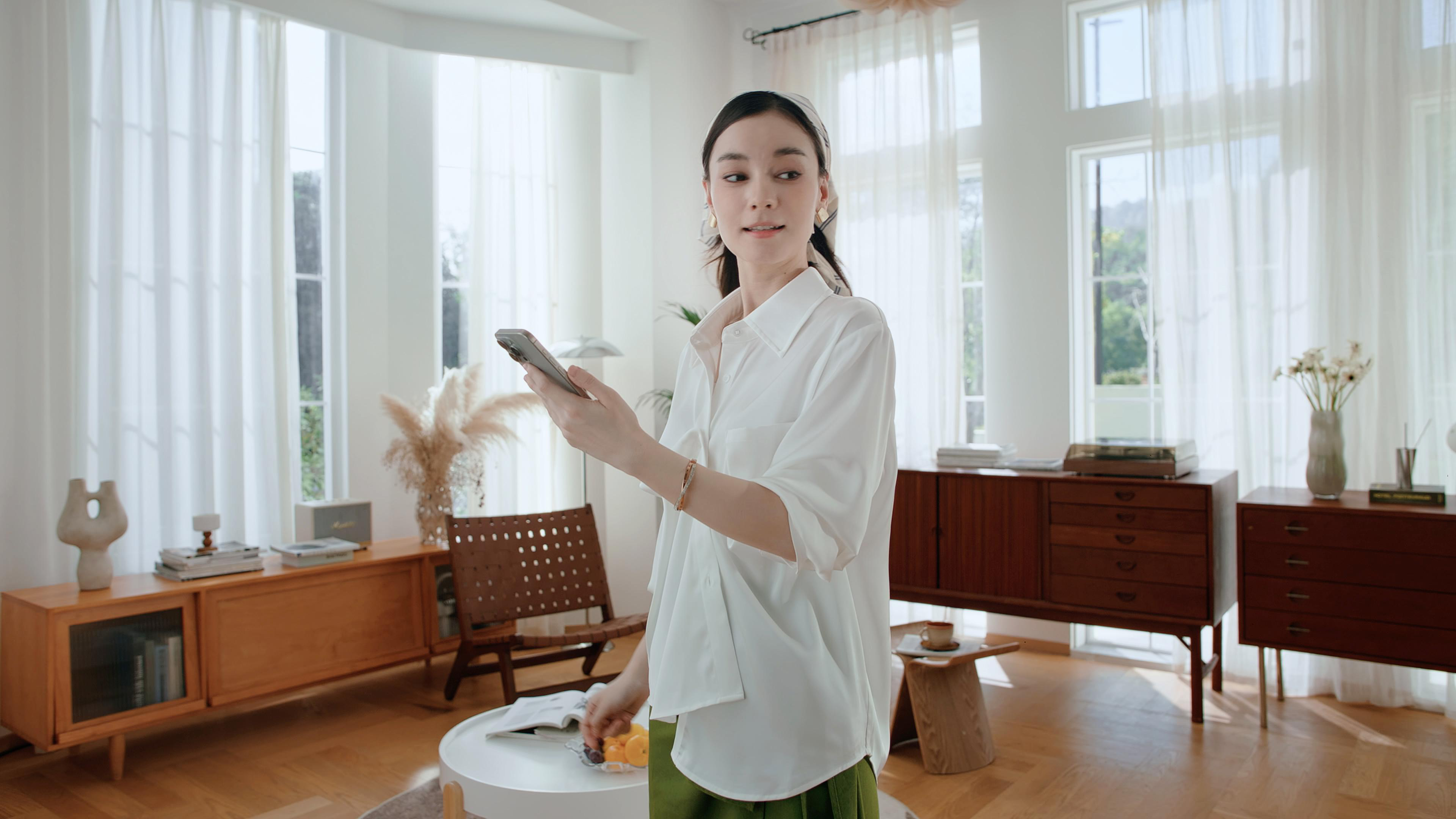 Eine Frau in einem weißen Hemd hält ein Handy in einem hellen Wohnzimmer.