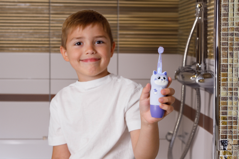 Chłopiec w białej koszulce trzyma fioletową szczoteczkę do zębów w kształcie kota.