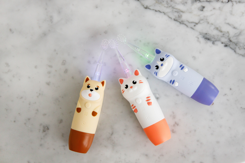 Trzy kolorowe szczoteczki do zębów z pyszczkami zwierząt i świecącymi główkami leżą na marmurze.