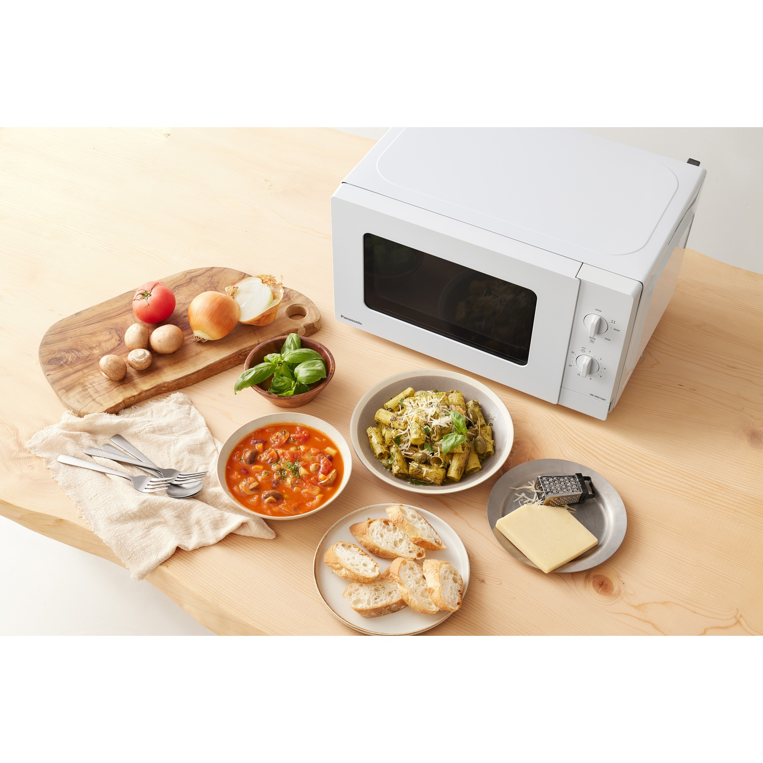 Un microonde bianco su un tavolo di legno con pasta, zuppa, pane e formaggio.