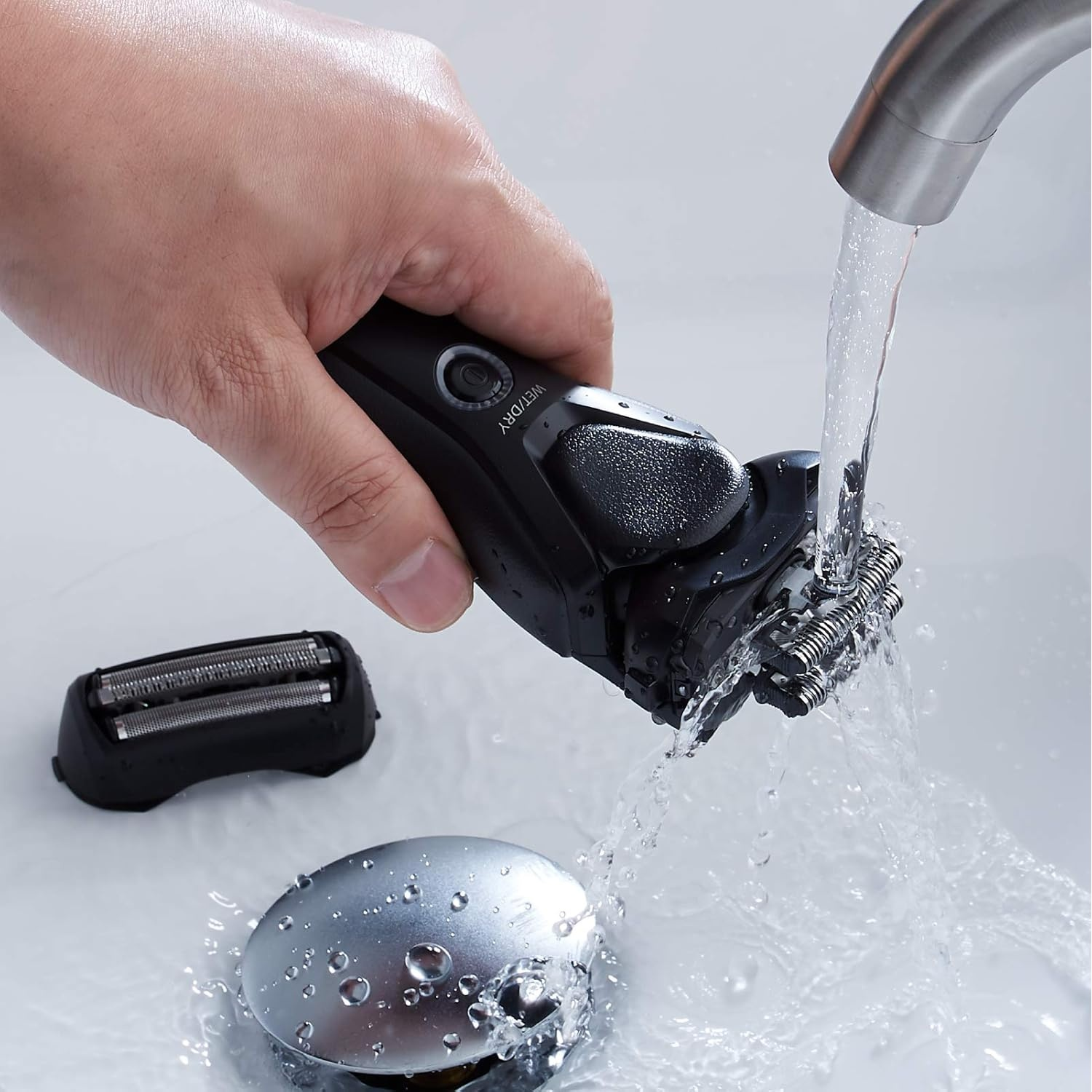 Una mano sciacqua un rasoio elettrico nero sotto l'acqua corrente in un lavandino.