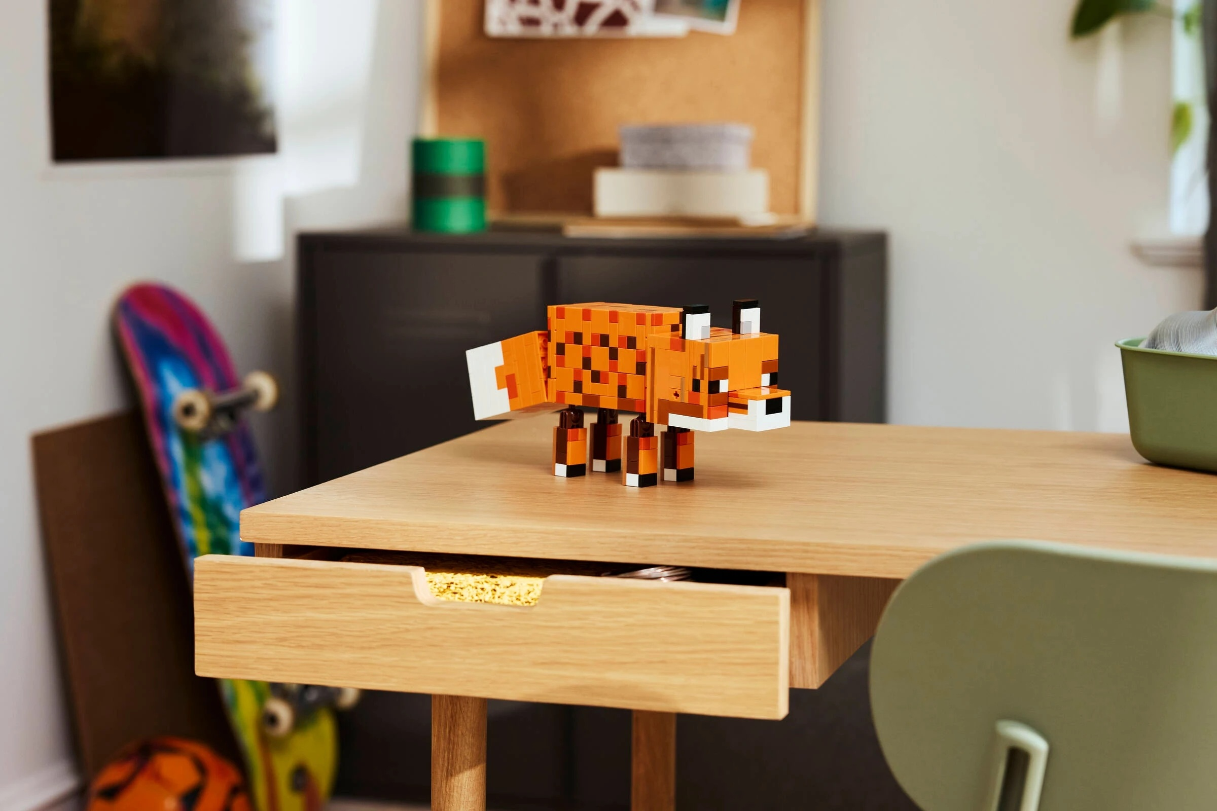 Pomarańczowy lis z klocków Lego stoi na drewnianym biurku. Deskorolka oparta o ścianę.