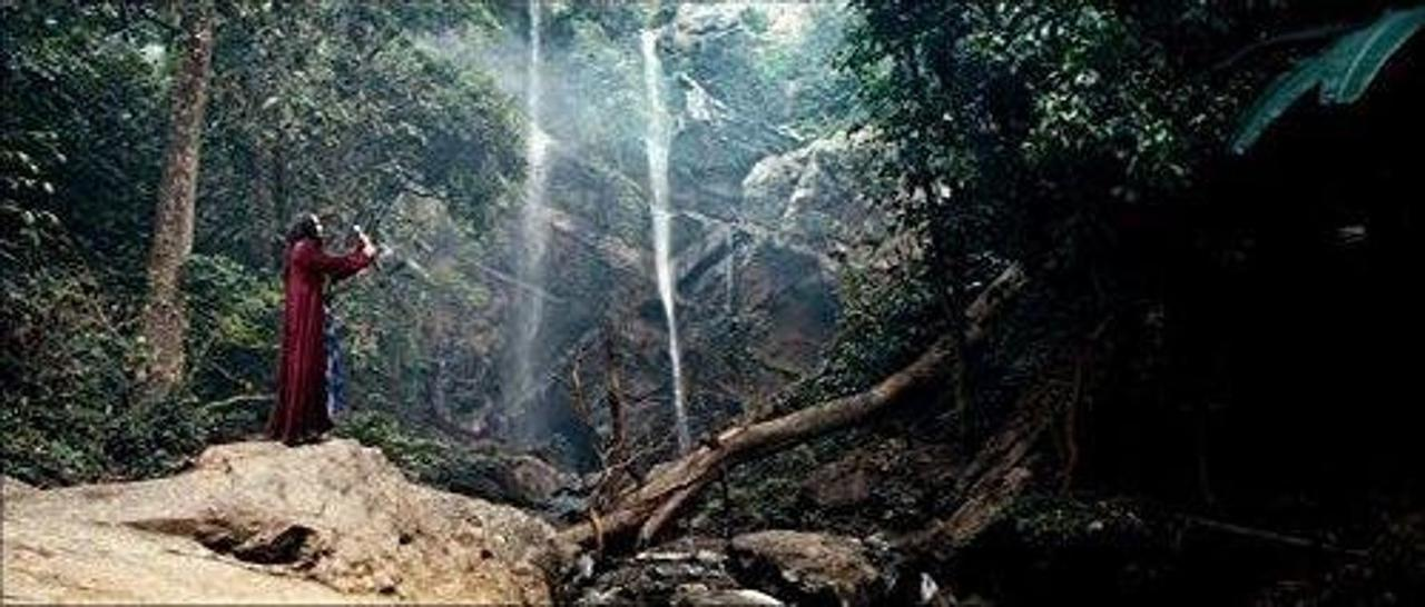 Eine Person in einem roten Gewand steht auf einem Felsen vor einem Wasserfall im Dschungel.