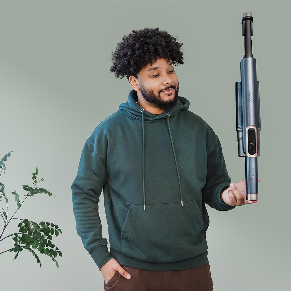 Un hombre con pelo afro sostiene una aspiradora azul de mano sobre un fondo verde claro.