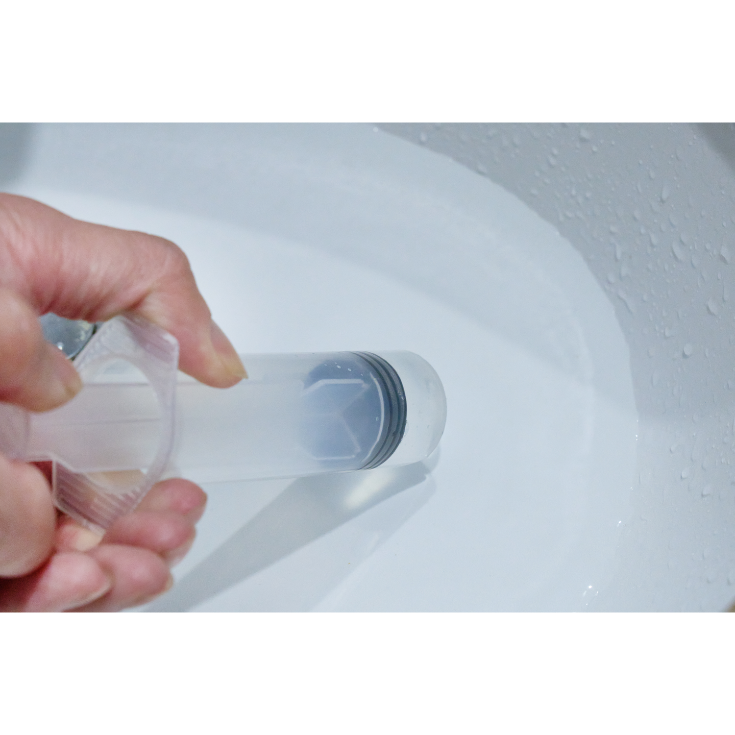 Een hand houdt een spuit boven een witte kuip met waterdruppels.