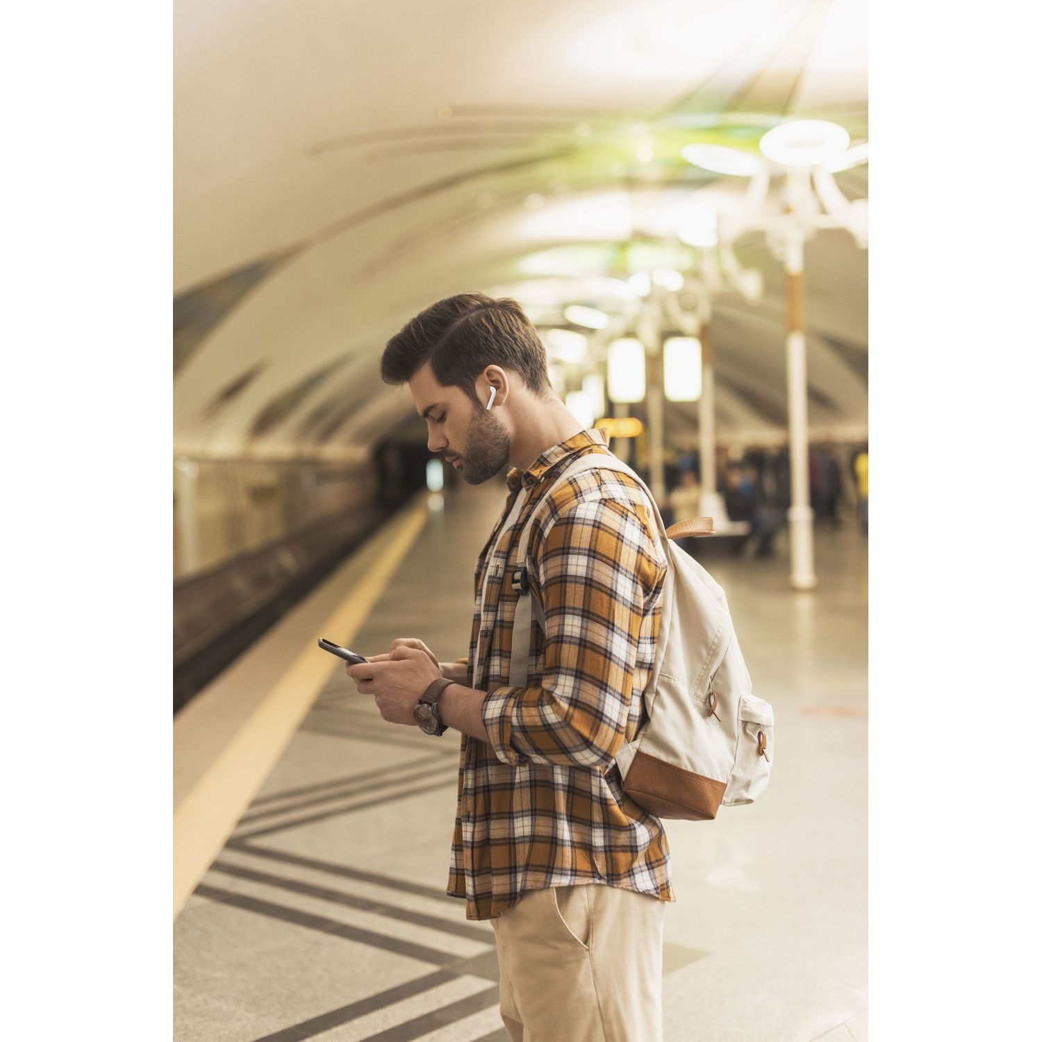 Uomo con zaino e auricolari su banchina metro guarda il telefono.