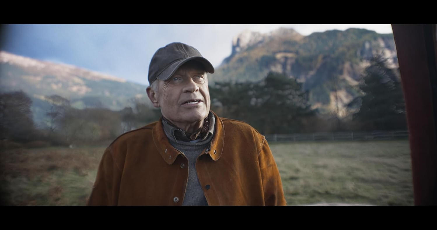 Ein älterer Mann mit Kappe und brauner Jacke steht vor einer bergigen Landschaft.