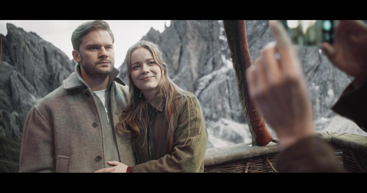 Ein Mann und eine Frau posieren in einem Heißluftballonkorb vor einer Berglandschaft.