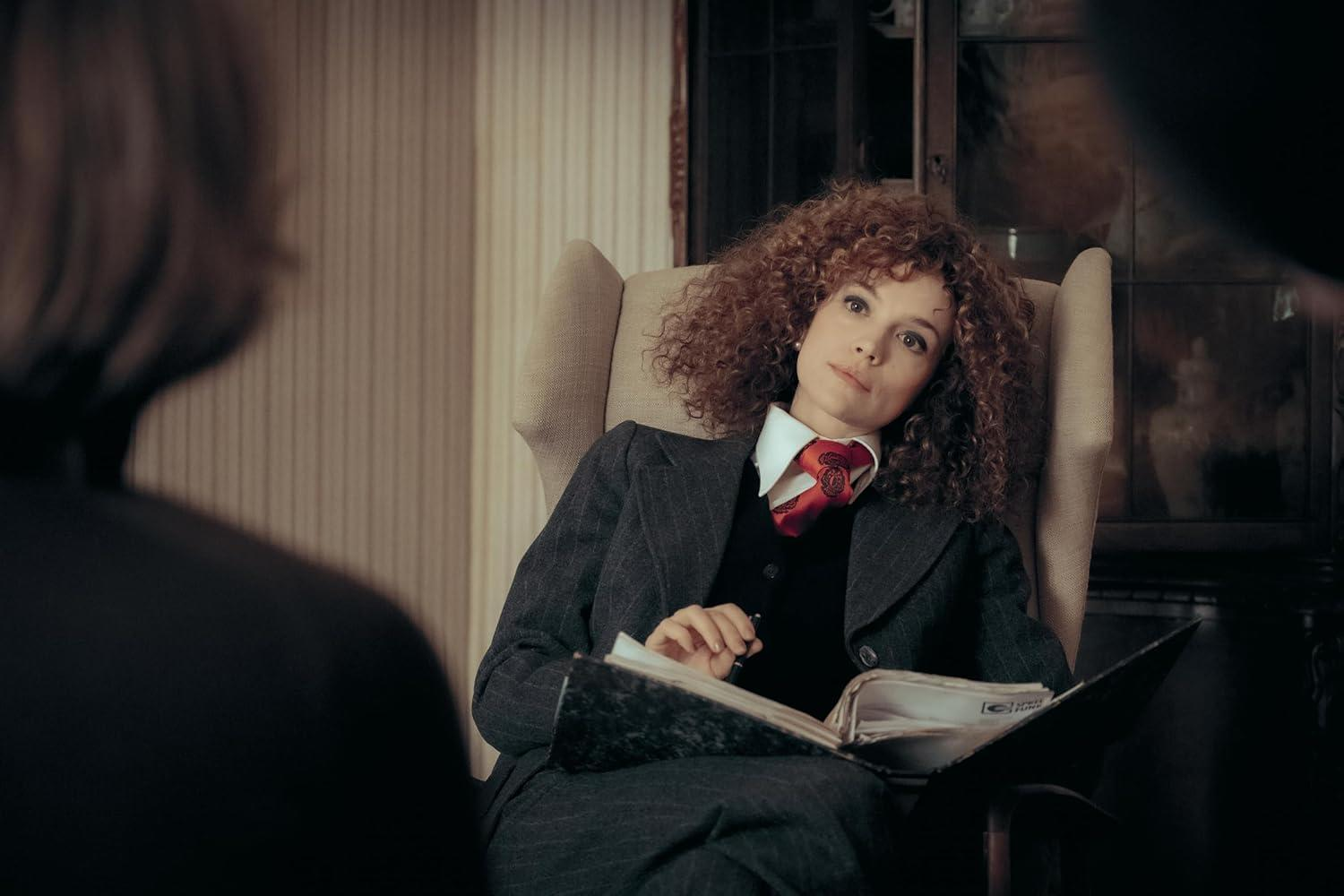 Frau mit roten Locken sitzt in einem Sessel, trägt einen Anzug und liest in einem Buch.