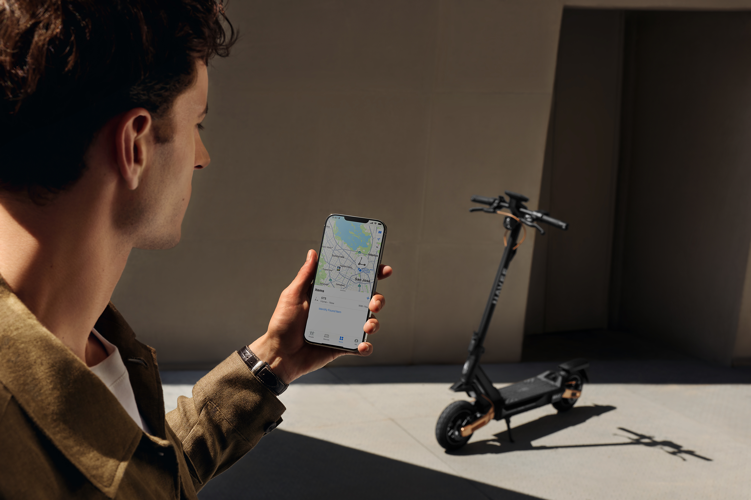 Ein Mann hält ein Smartphone mit einer Karte. Ein E-Scooter steht im Hintergrund.