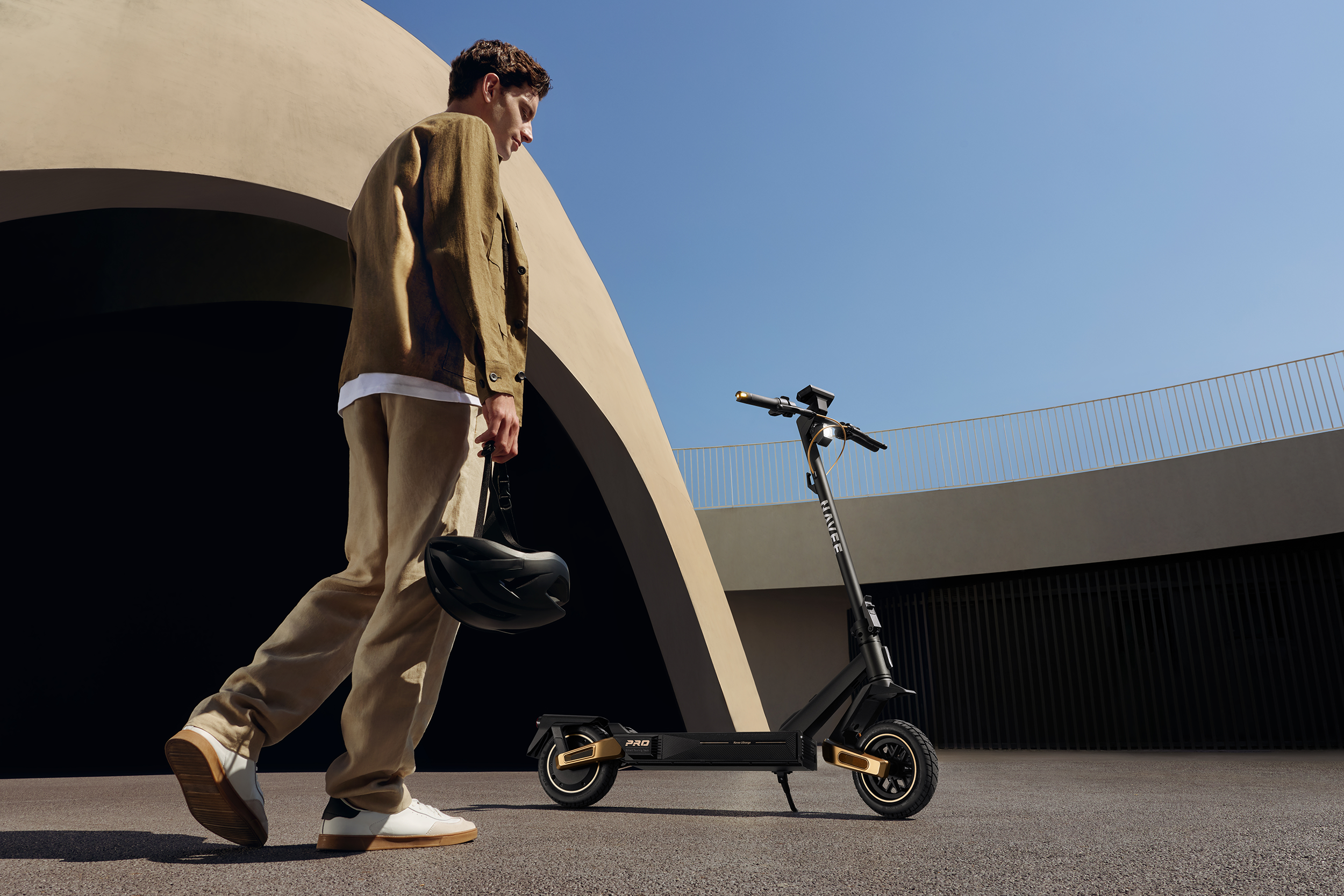 Ein Mann mit brauner Jacke trägt einen schwarzen Helm und geht neben einem schwarzen E-Scooter.
