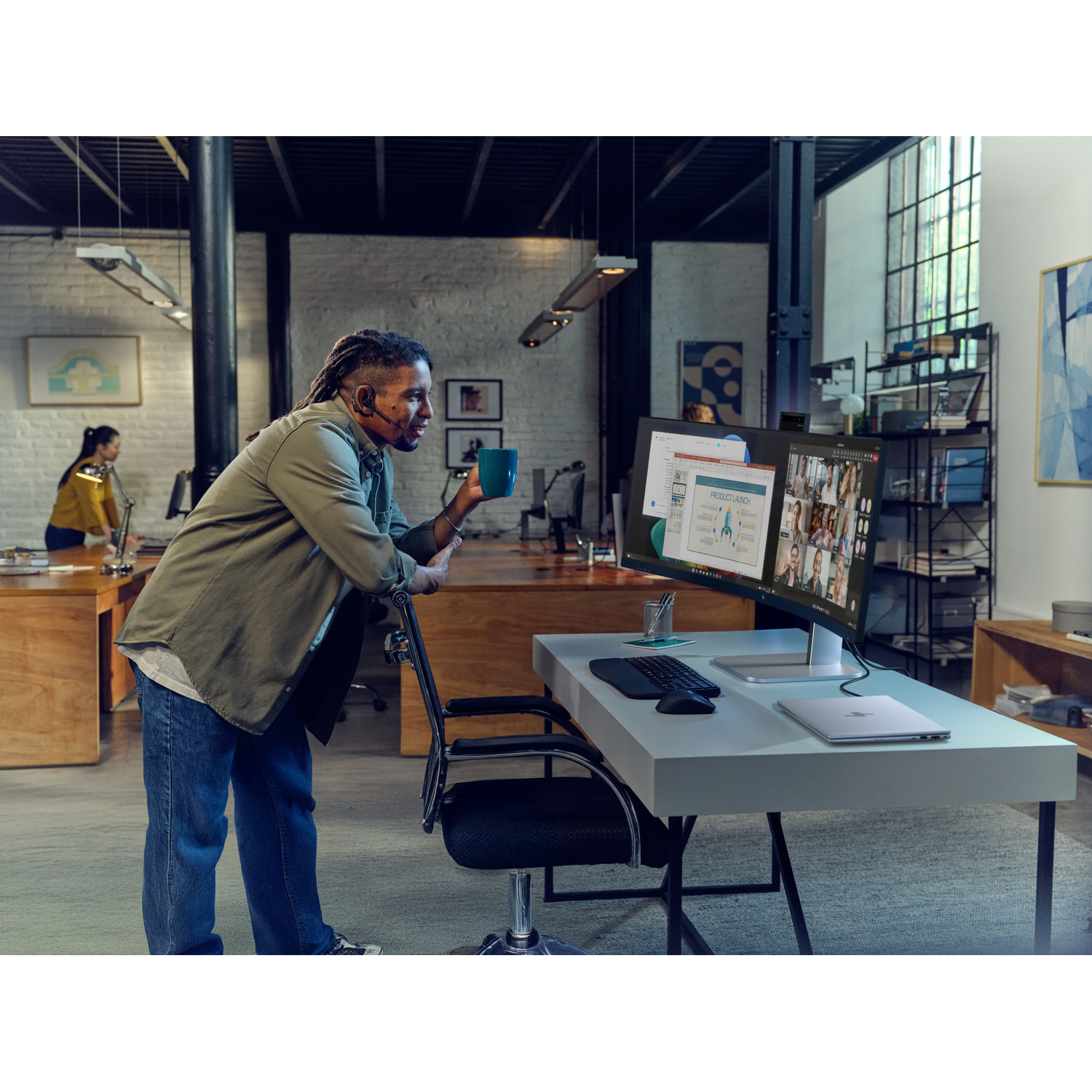 Man with dreadlocks holds mug next to dual monitors showing video conference.