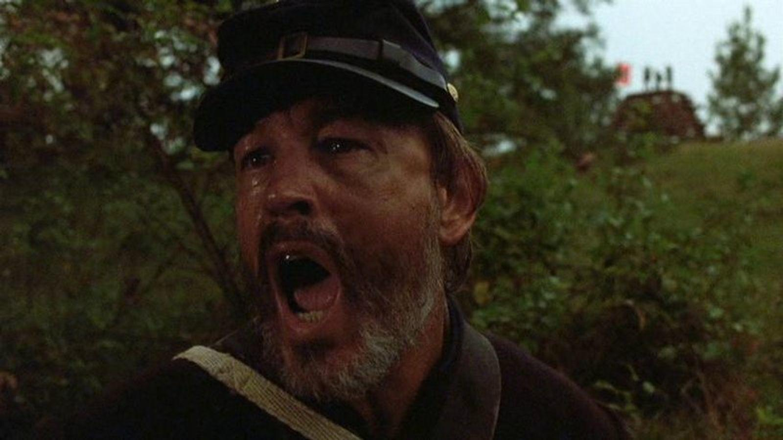 Ein Mann in Uniform mit offenem Mund und Tränen in den Augen, im Hintergrund Bäume.