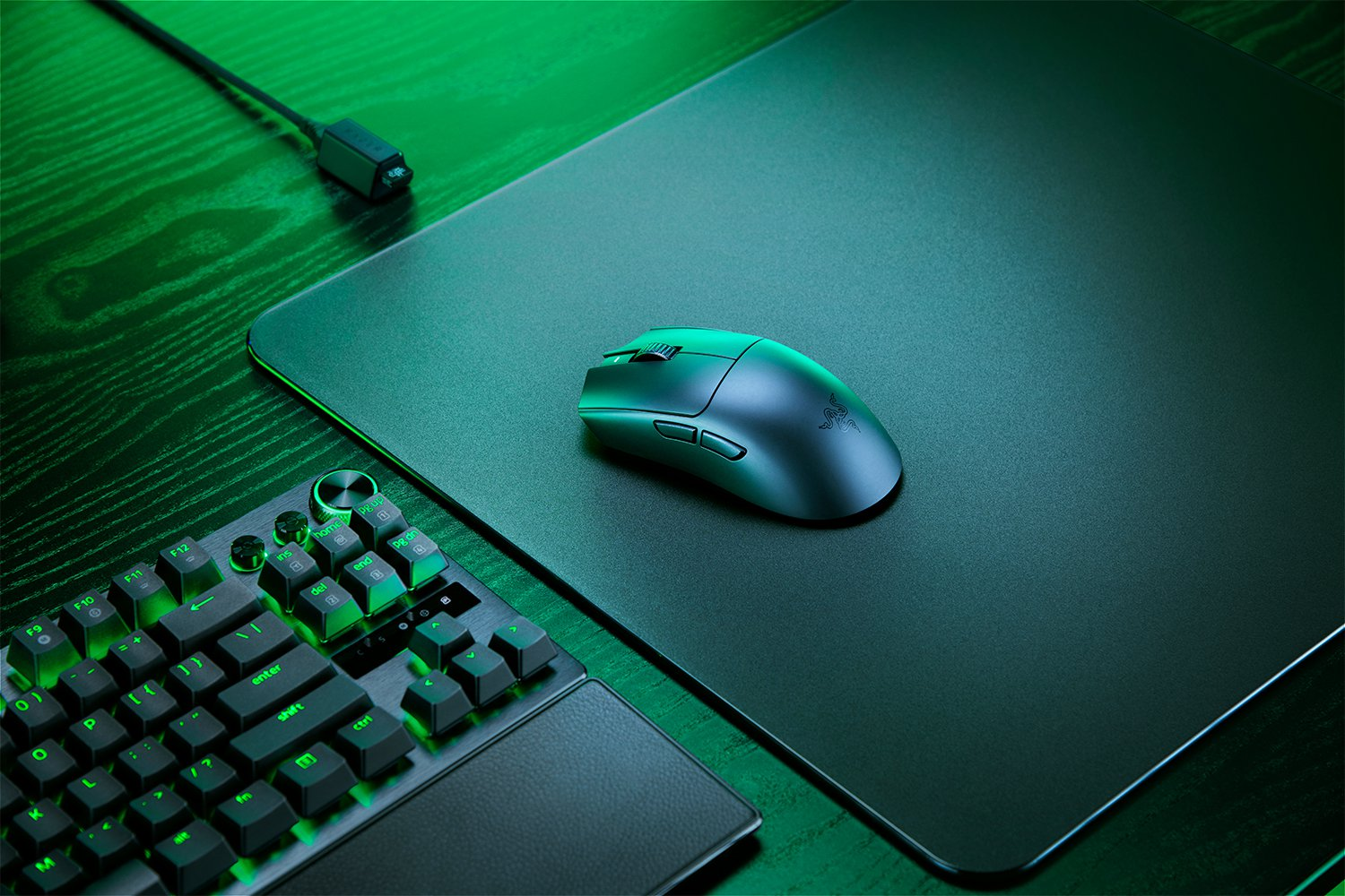 Un ratón y teclado gaming oscuros con retroiluminación verde sobre una alfombrilla negra.