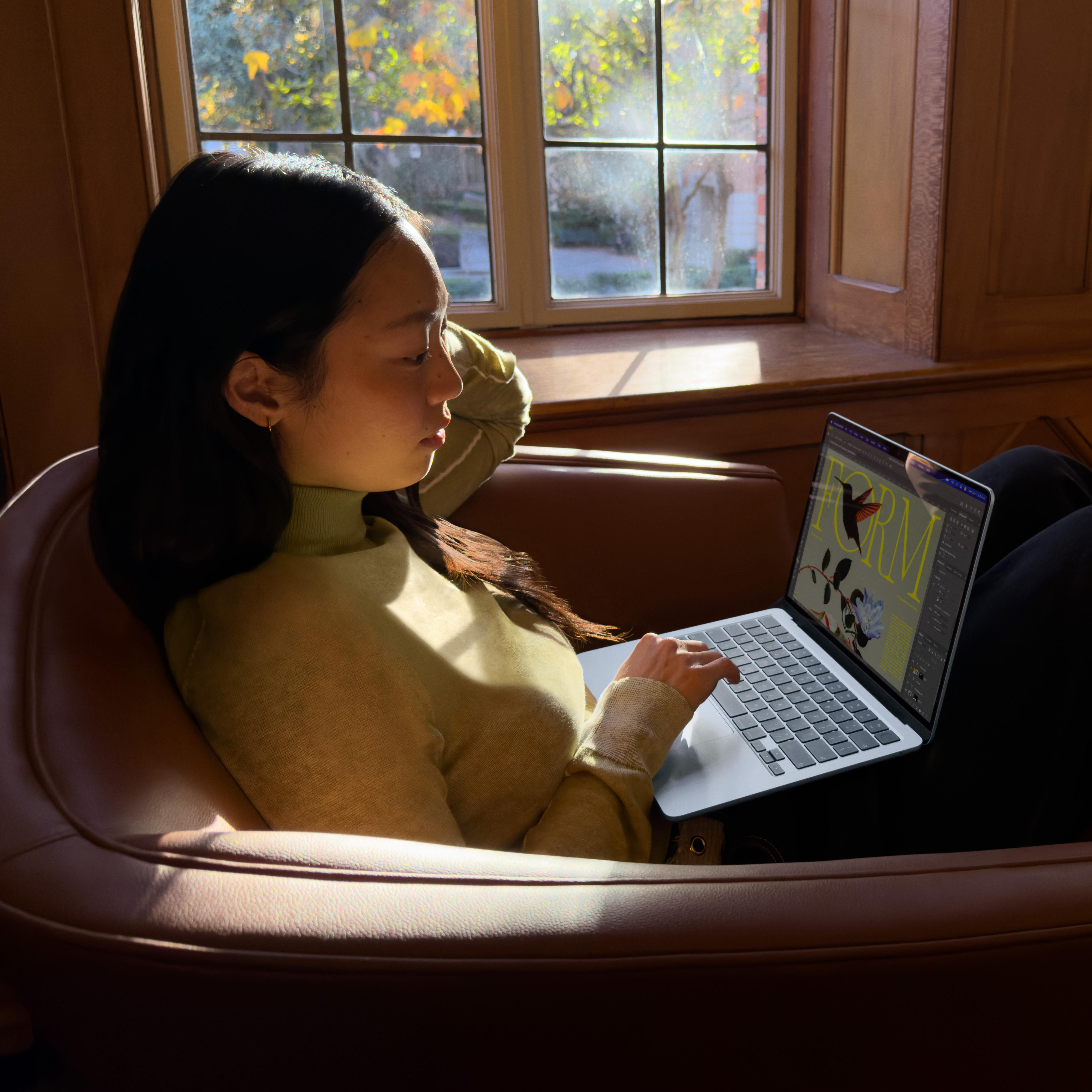 Eine Frau sitzt in einem braunen Sessel und arbeitet an einem Laptop neben einem Fenster.