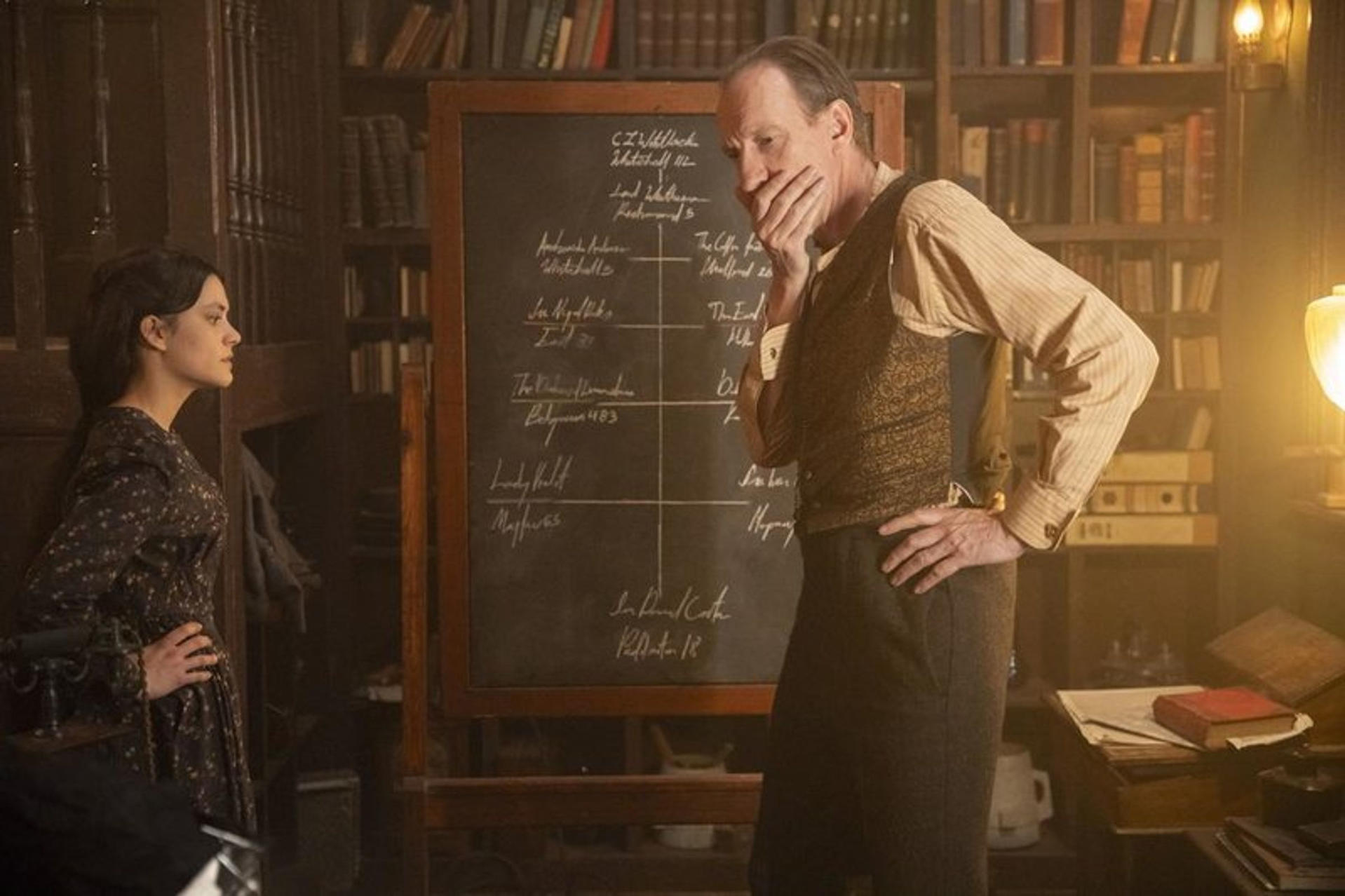Ein Mann in einer Weste und eine Frau in einem geblümten Kleid stehen in der Nähe einer Tafel in einer Bibliothek.