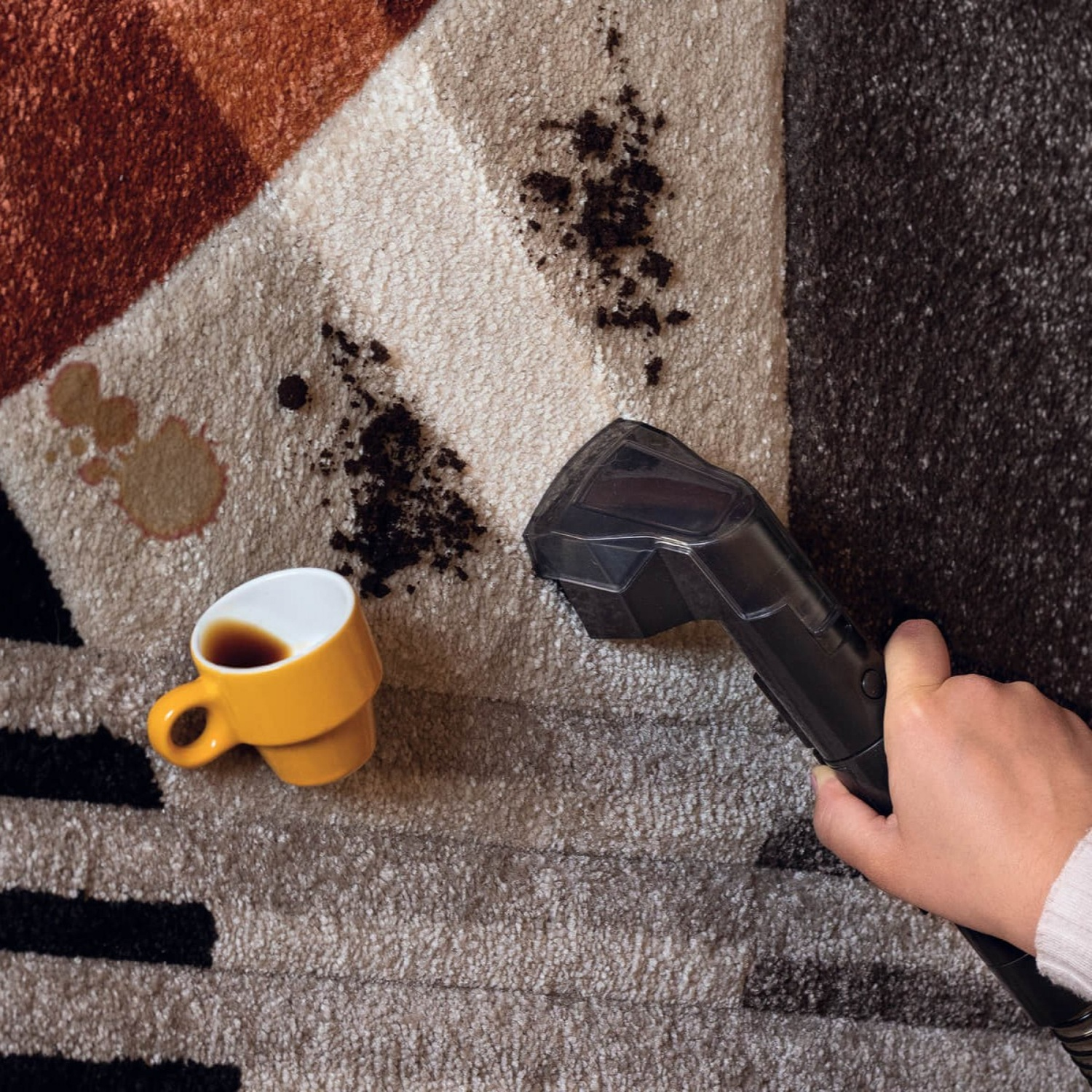 Una mano aspira sporco da un tappeto fantasia vicino a una tazza di caffè rovesciata.