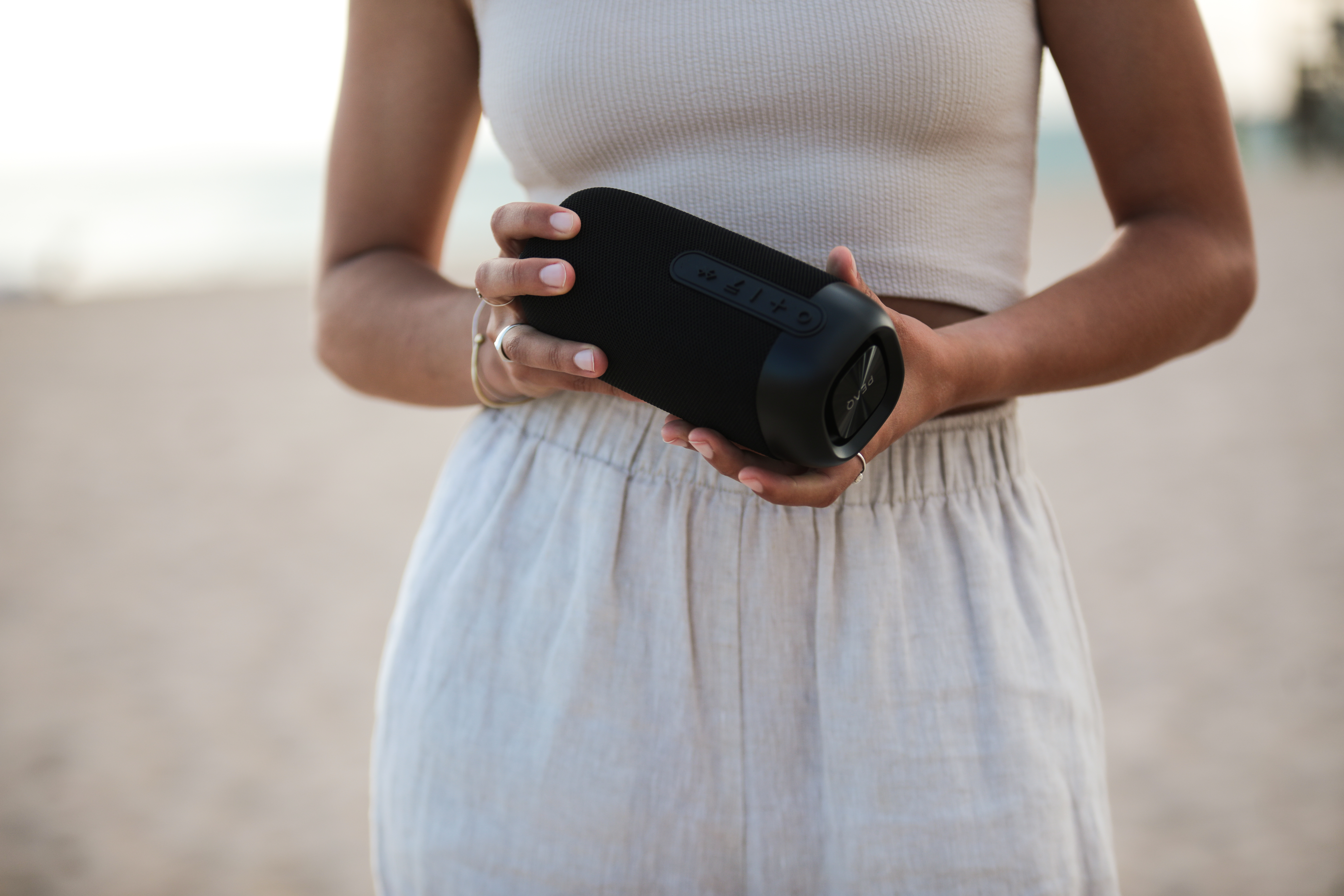 Kobieta trzyma czarny głośnik Bluetooth na plaży, jasne ubranie.