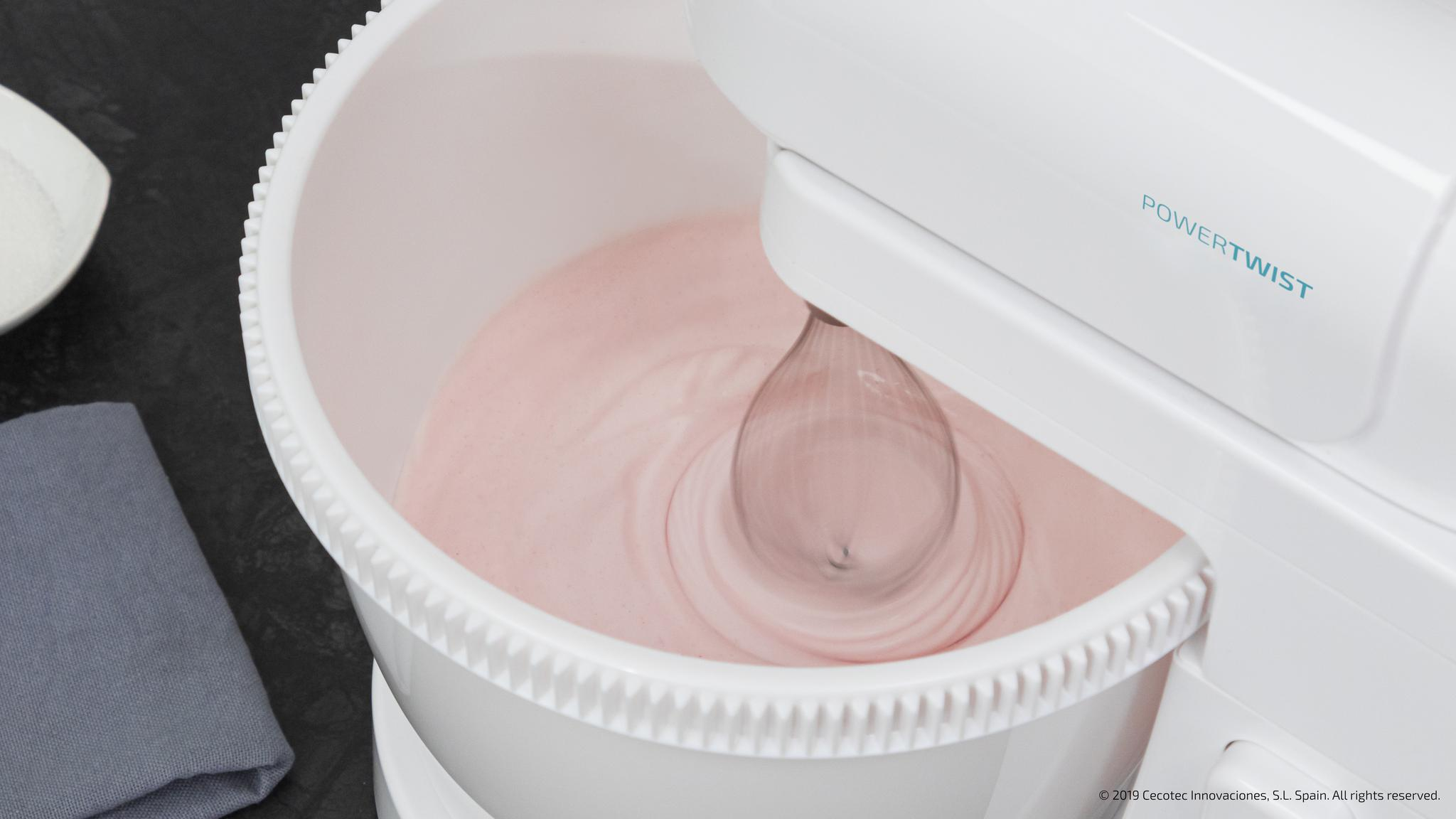 Una batidora blanca bate crema rosa en un bol. Un paño gris está cerca.