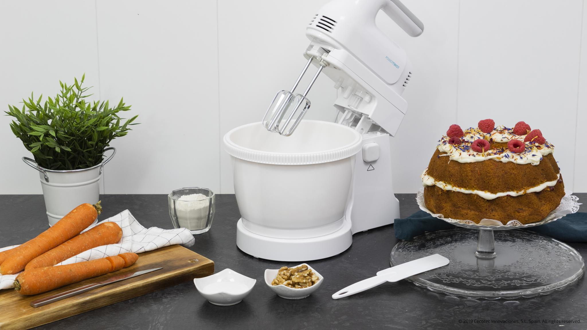 Un batidor de mano blanco con bol, pastel, zanahorias y nueces cerca.