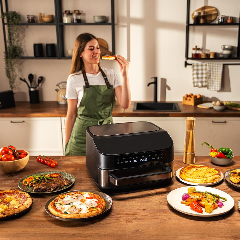 Una mujer come pizza en una cocina con una freidora de aire y comida en la mesa.