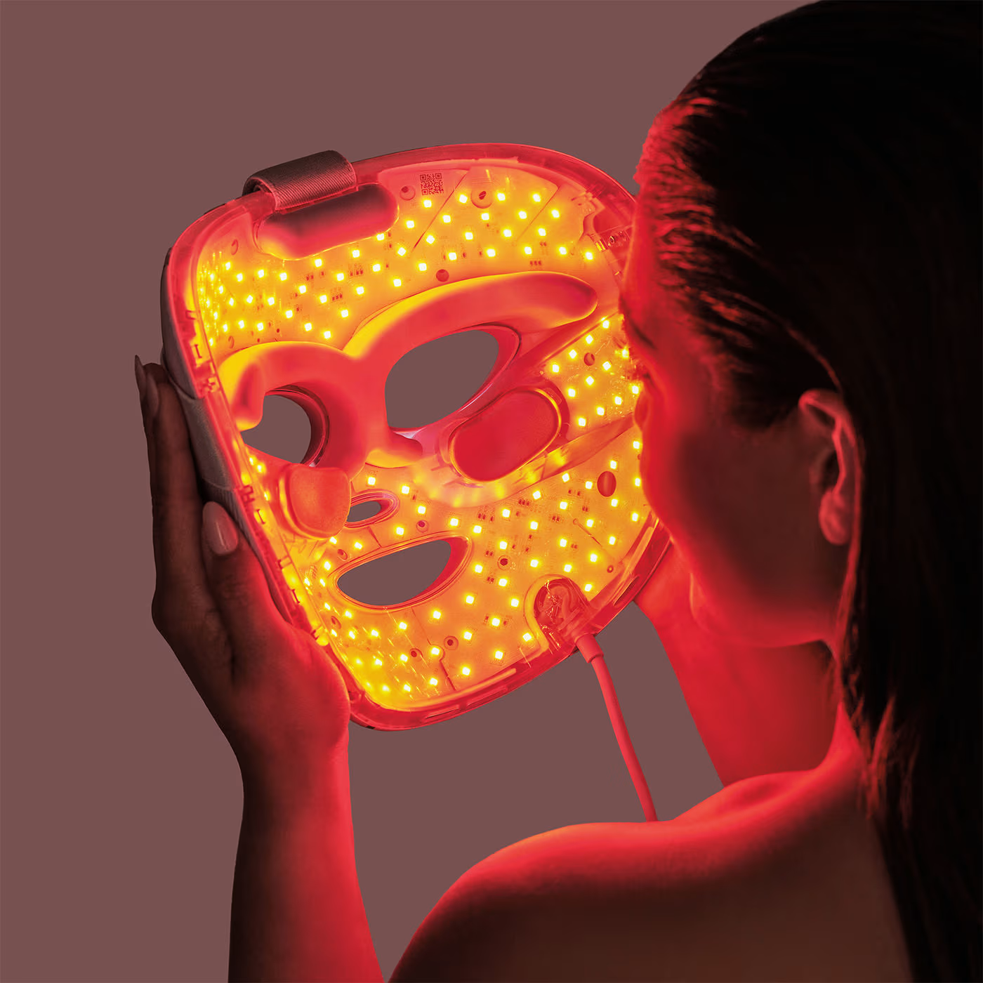 Une femme tient un masque facial LED avec des lumières rouges devant son visage.