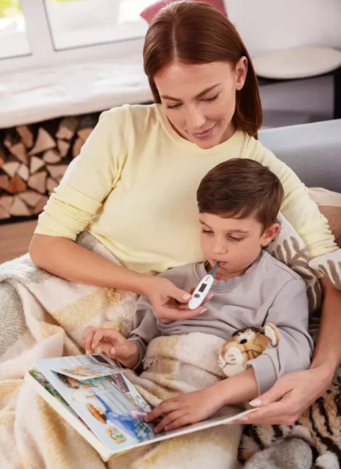 Kobieta mierzy dziecku temperaturę termometrem podczas wspólnego czytania książki.