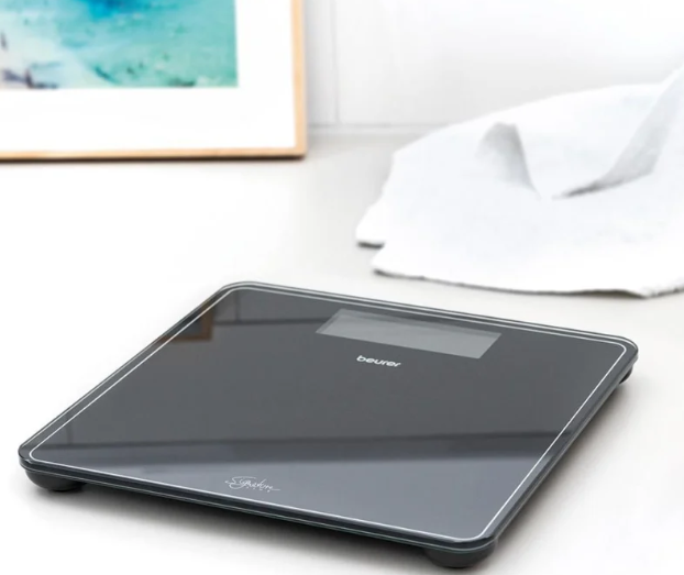 Nowoczesna czarna waga cyfrowa z wyświetlaczem stoi na białej powierzchni obok złożonego ręcznika.