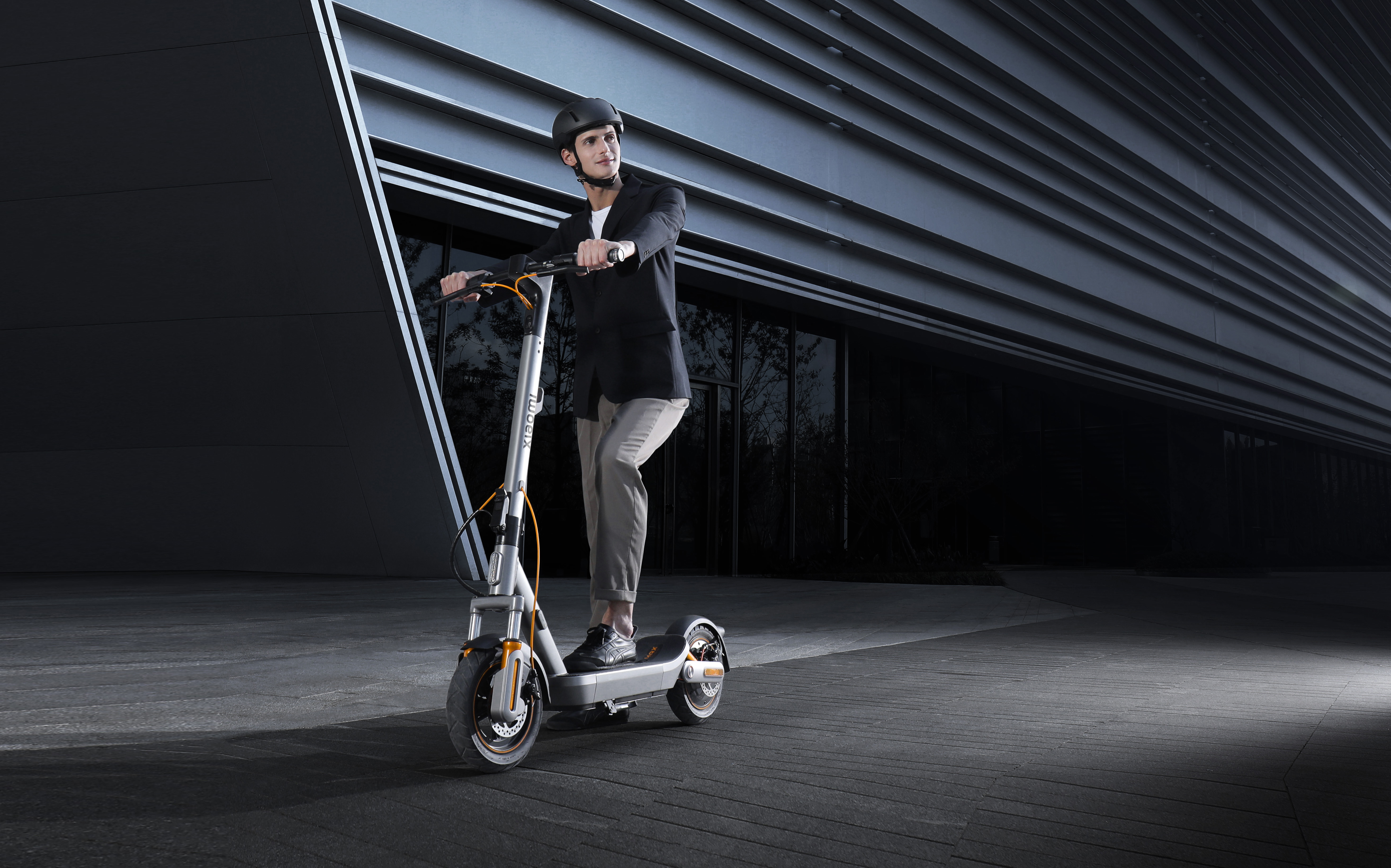 Mann in schwarzem Sakko und Helm fährt E-Scooter vor modernem Gebäude.