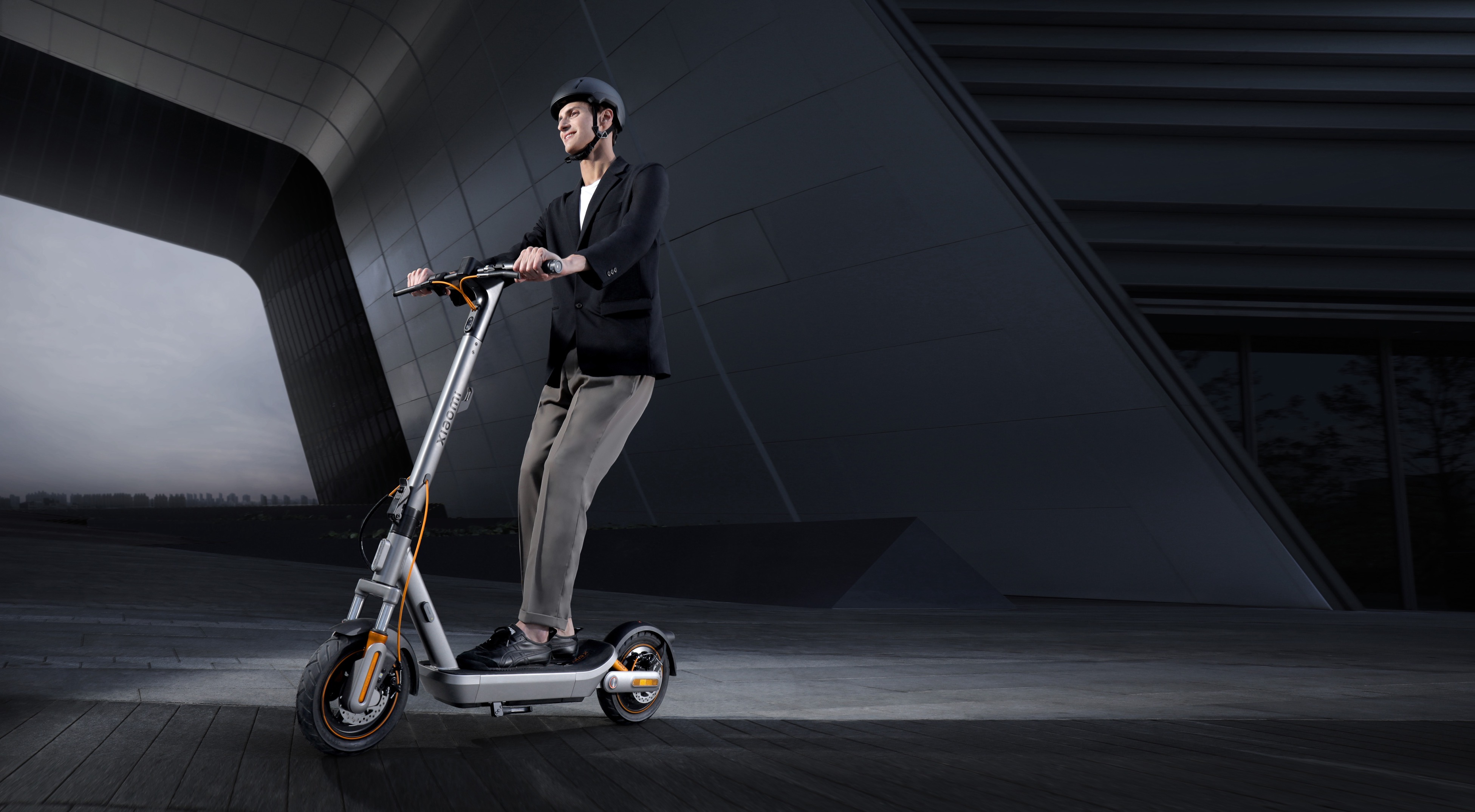Ein Mann in einem schwarzen Sakko fährt einen silbernen E-Scooter vor einem modernen Gebäude.