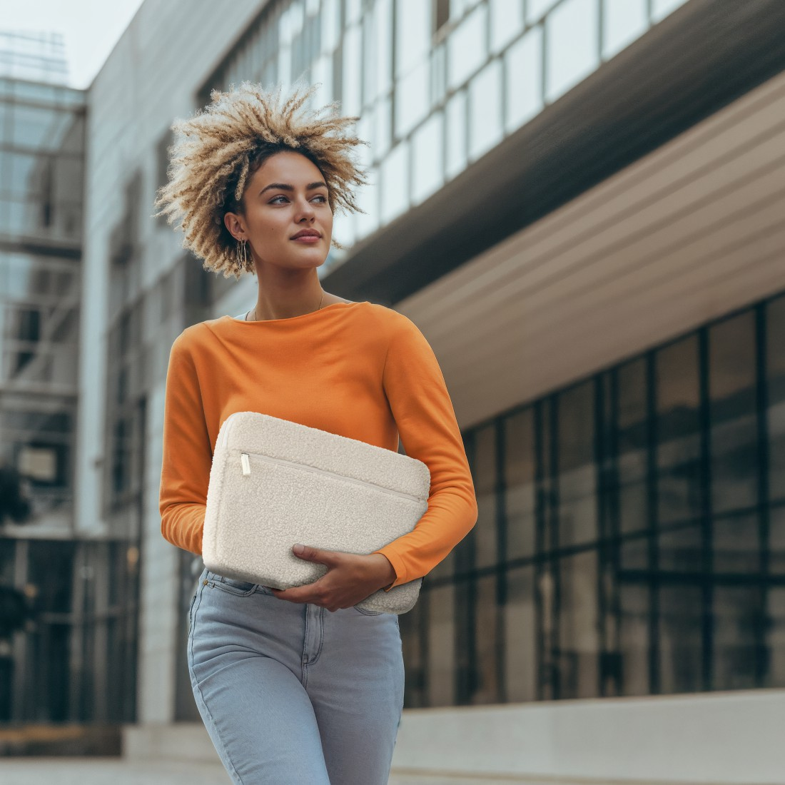 Eine Frau mit lockigem Haar trägt einen orangefarbenen Pullover und hält eine weiße Laptoptasche.