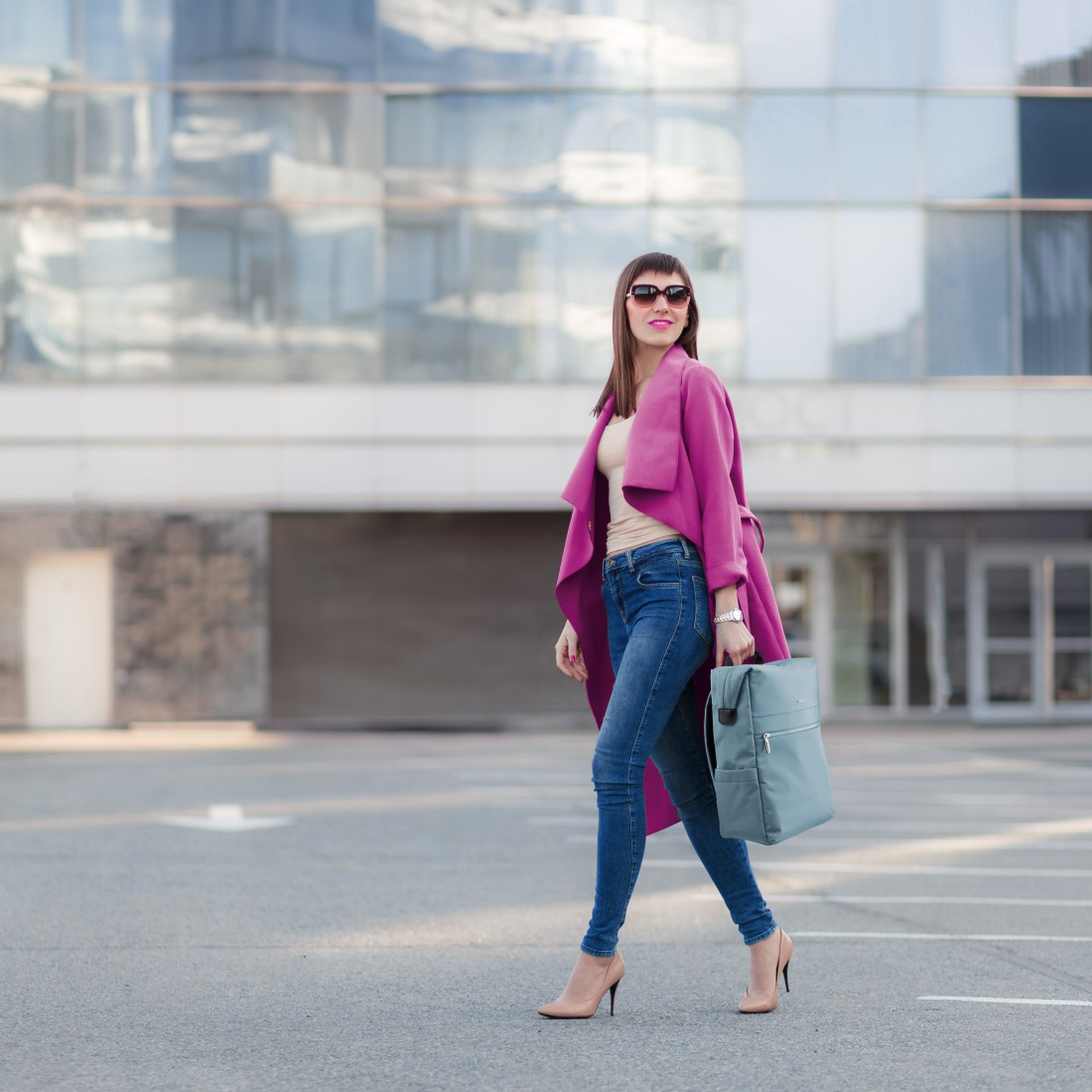 Frau in pinkem Mantel und Jeans geht mit Rucksack, moderne Glasfassade im Hintergrund.