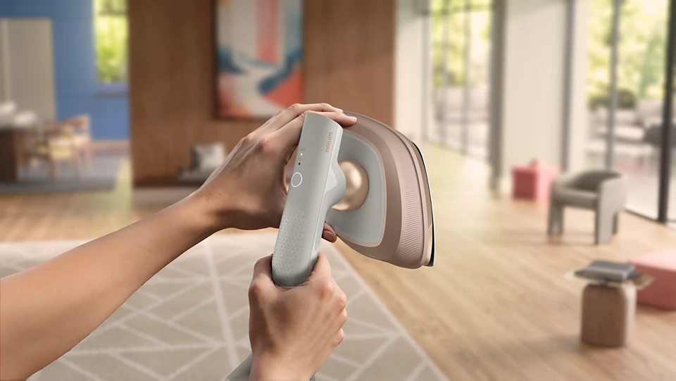 Dłonie trzymają jasnoszarą parownicę ręczną w kolorze różowego złota na wzorzystym dywanie.