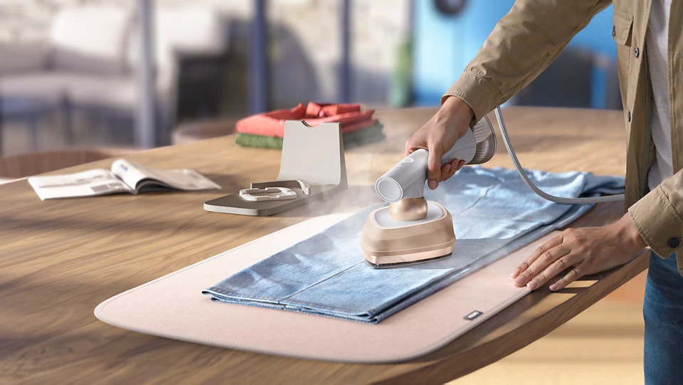 Dłoń używa ręcznej parownicy do niebieskiego ubrania na drewnianym stole.