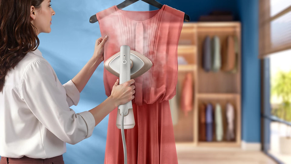 Kobieta prasuje parą różową sukienkę przed szafą. Widoczna para.